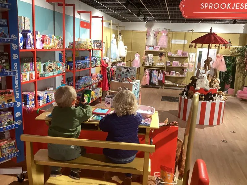 Twee peuters zitten op een bankje en spelen aan een tafeltje in de winkel. Op de achtergrond rode kasten met Lego, verkleedjurken en een rood-wit gestreepte carrousel met knuffels.